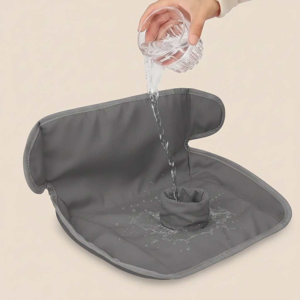 A grey waterproof floor mat or diaper cover with a central drain opening, shown with a hand pouring water from a clear glass to demonstrate its water-resistant, draining design. The background is a solid, warm beige color.